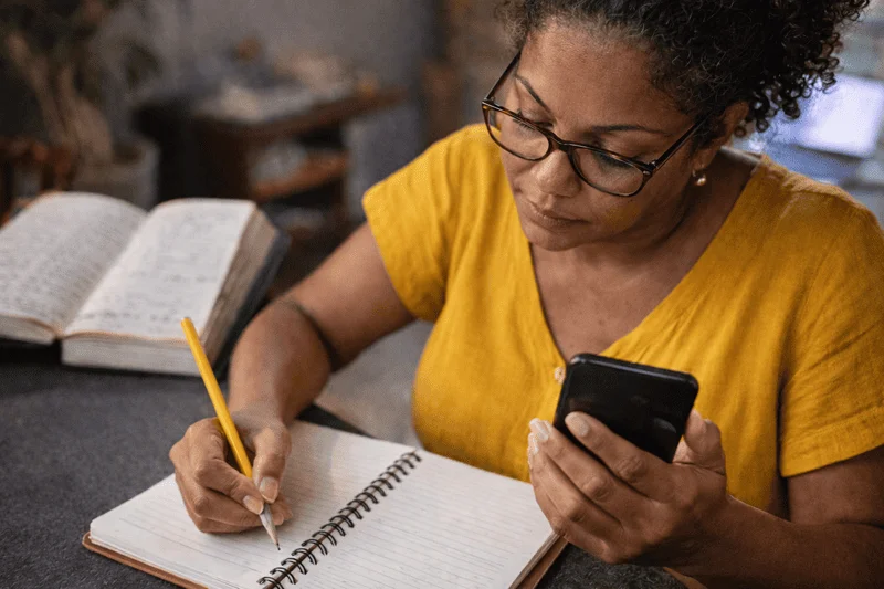 Pessoa segurando celular e escrevendo com lápis em um caderno, representando a evolução do crédito no Brasil do caderno da venda ao digital